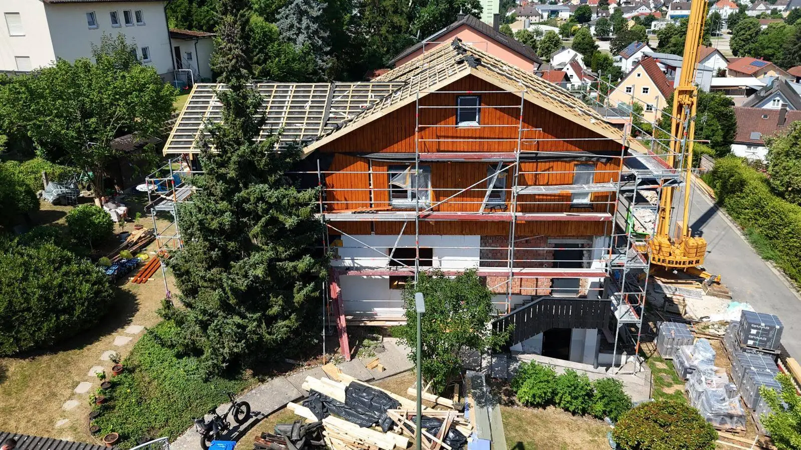 Single-family house under construction: gable end with new timber façade, open roof truss and scaffolding, crane on the street side